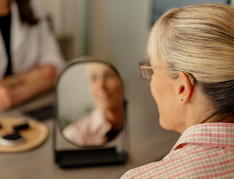 Woman being fit with hearing aids smiling into a mirror.
